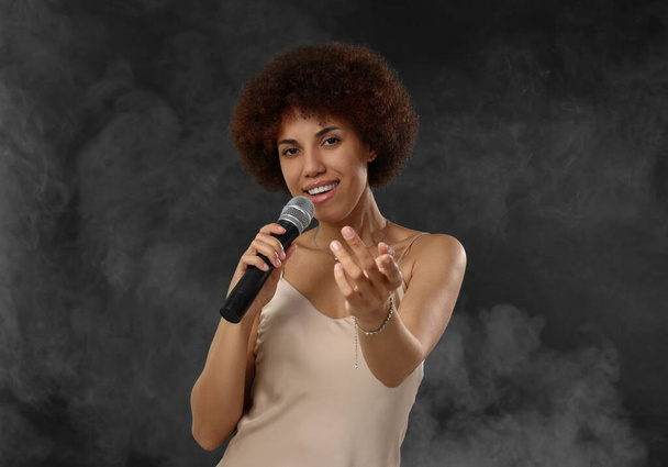 krullend jong vrouw met microfoon zingen in rook op zwart achtergrond - Foto, afbeelding