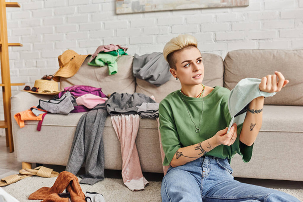 stylish tattooed woman holding cap and looking away while sitting on floor near couch with clothes, wardrobe items sorting, sustainable living and mindful consumerism concept - Photo, Image