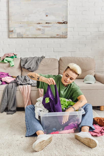 stylish tattooed woman with trendy hairstyle sorting clothes in plastic container on floor near couch with wardrobe items in modern living room, sustainable living and mindful consumerism concept - Photo, Image
