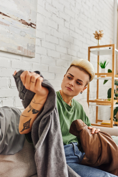 mujer tatuada con peinado de moda clasificación de ropa y la reducción de artículos de armario en el sofá en la sala de estar cerca de rack con plantas verdes en el hogar, vida sostenible y concepto de consumismo consciente - Foto, imagen