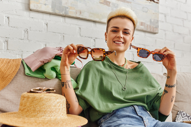 happy young woman sitting on couch near straw hat and clothing while showing stylish sunglasses, trendy hairstyle, tattoo, sustainable living and mindful consumerism concept - Photo, Image