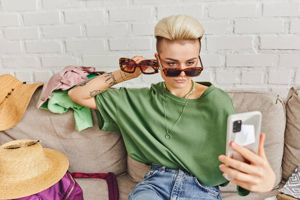 femme tatouée élégante avec des lunettes de soleil prenant selfie sur smartphone près de chapeaux de paille et vêtements sur canapé, échange en ligne, marché virtuel, vie durable et concept de consumérisme conscient - Photo, image
