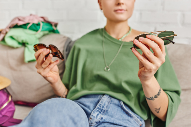 partial view of casual style and tattooed woman holding trendy sunglasses near clothing on couch, decluttering, sorting, sustainable living and mindful consumerism concept - Photo, Image