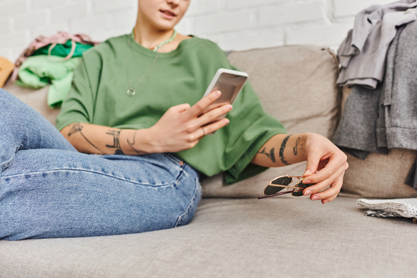 rajattu näkymä nuoresta ja tatuoidusta naisesta, joka istuu sohvalla kotona ja ottaa valokuvan aurinkolaseista älypuhelimella online-vaihtoa, kestävää elämää ja tietoista kulutuskonseptia varten - Valokuva, kuva