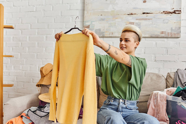 młoda wytatuowana kobieta uśmiechnięta i patrząca na żółty modny sweter siedząca na kanapie w pobliżu sklepu z używanymi rzeczami znajduje się w nowoczesnym salonie w domu, zrównoważone życie i świadoma koncepcja konsumpcjonizmu - Zdjęcie, obraz