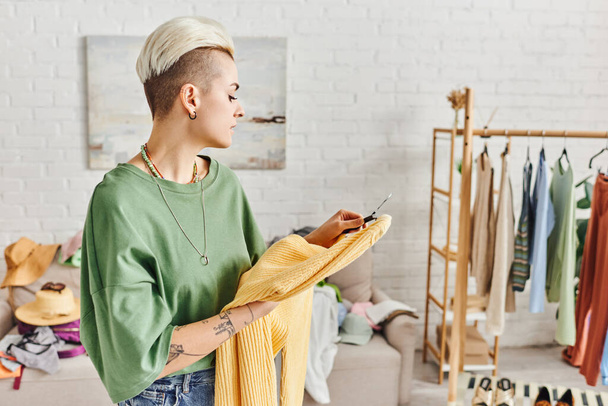 stylová tetovaná žena při pohledu na žlutý svetr v blízkosti stojanu s oblečením a obuví v moderním obývacím pokoji, třídění oblečení, udržitelné bydlení a bdělý konzumní koncept - Fotografie, Obrázek