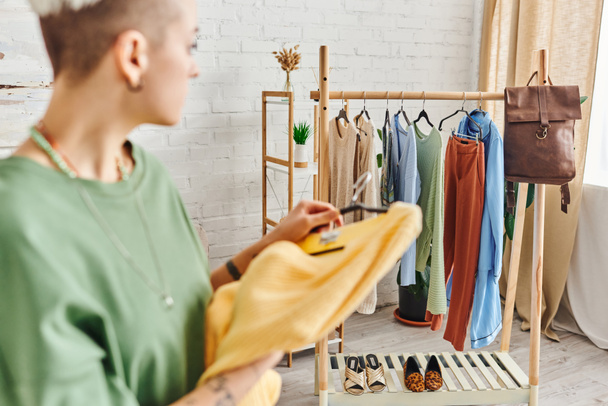 zamazana kobieta z żółtym swetrem patrząca na stojak z wieszakami, ubraniami, skórzaną torbą i obuwiem w nowoczesnym salonie, sortowanie elementów garderoby, zrównoważone życie i świadoma koncepcja konsumpcjonizmu - Zdjęcie, obraz