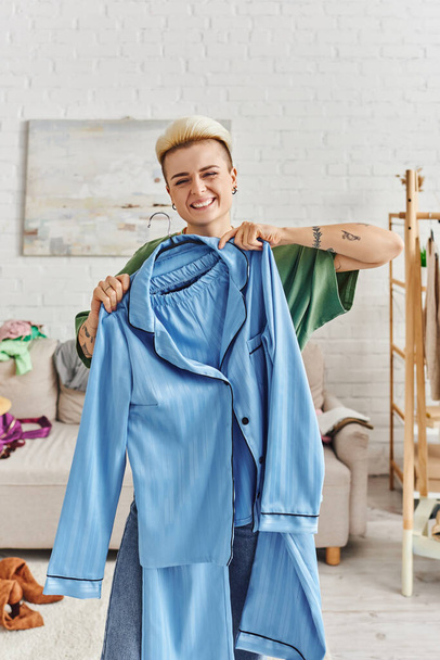 mujer tatuada llena de alegría con un peinado de moda que muestra pijamas azules mientras clasifica la ropa y reduce el armario en la sala de estar en casa, la vida sostenible y el concepto de consumismo consciente - Foto, Imagen