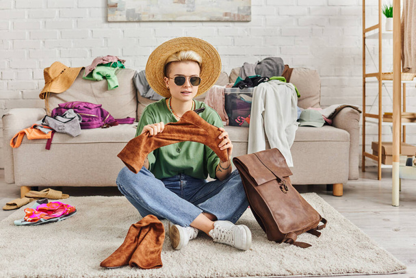 Sparsame Kleidung, modische Frau mit Strohhut und Sonnenbrille, die auf dem Boden neben der Ledertasche sitzt und Wildlederstiefel in der Nähe der Couch im Wohnzimmer hält, nachhaltige Mode und bewusstes Konsumkonzept - Foto, Bild
