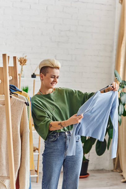 wytatuowana i zadowolona kobieta przyglądająca się niebieskiemu sweterkowi podczas sortowania ubrań w pobliżu regału z elementami garderoby na wieszakach w salonie w domu, zrównoważona moda i świadoma koncepcja konsumpcjonizmu - Zdjęcie, obraz