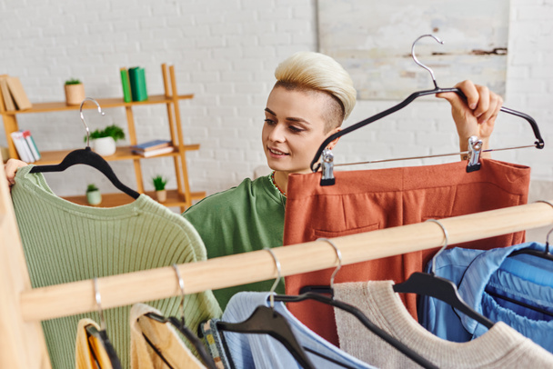positive young woman holding hangers with pants and jumper near rack with wardrobe garments, clothing sorting, conscious decluttering, sustainable fashion and mindful consumerism concept - Photo, Image
