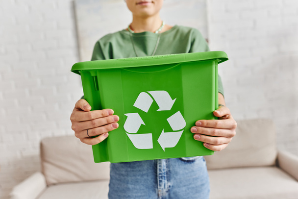 focus on green plastic box with recycling sign in hands of cropped woman standing at home on blurred background, sustainable living and environmentally friendly habits concept - Photo, Image