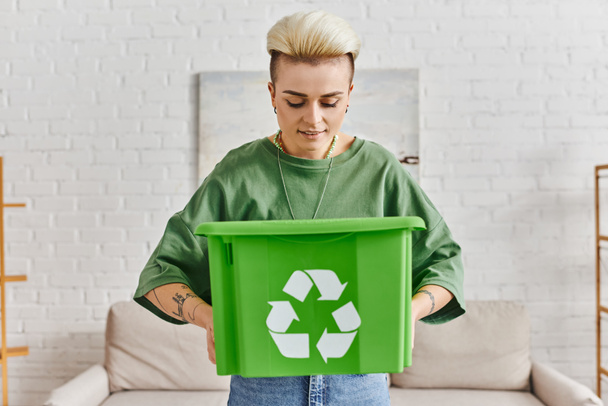 uśmiechnięta, wytatuowana kobieta z modną fryzurą, wyglądająca w zielonym plastikowym pudełku ze znakiem recyklingu, stojąca w domu, redukująca marnotrawstwo, zrównoważone życie i przyjazne środowisku nawyki - Zdjęcie, obraz