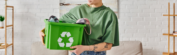 vista parcial de la joven mujer tatuada en ropa casual que sostiene la papelera de reciclaje verde con prendas de vestir en la sala de estar moderna, concepto de hábitos sostenibles y respetuosos con el medio ambiente, bandera - Foto, Imagen