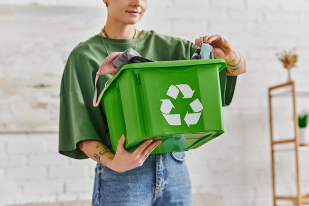 partial view of tattooed woman holding green recycling box with clothes while standing in living room, ethical consumption, sustainable living and environmentally friendly habits concept - Photo, Image