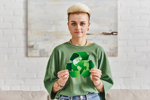 pozitív tetovált nő trendi frizurával mutatja zöld újrahasznosítási jel földgömbbel, miközben mosolyog a kamera modern nappaliban, fenntartható életmód és a környezettudatosság koncepció - Fotó, kép