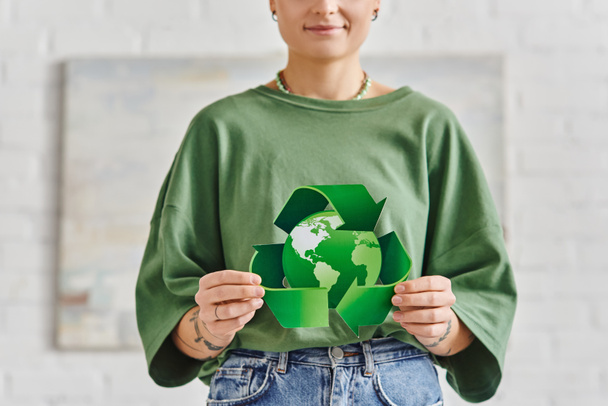 ekotietoinen elämäntapa, osittainen näkemys hymyilevästä tatuoidusta naisesta rennoissa vaatteissa, joilla on vihreä kierrätyssymboli ympäri maailmaa kotona, kestävä elin- ja ympäristötietoisuus - Valokuva, kuva