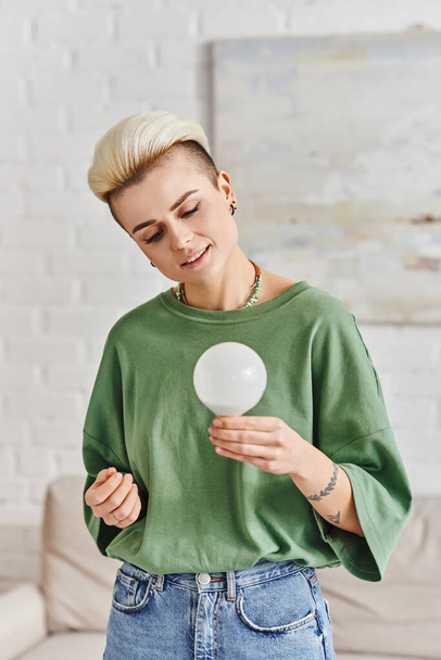 green living, young tattooed woman with trendy hairstyle and happy face holding energy saving light bulb at home, sustainable lifestyle and environmentally conscious concept - Foto, imagen