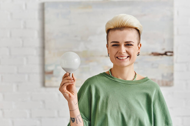 eficiência ecológica, mulher tatuada alegre com penteado moderno segurando lâmpada economizadora de energia e olhando para a câmera na sala de estar, estilo de vida sustentável e conceito ambientalmente consciente - Foto, Imagem
