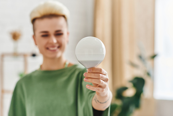 energy saving light bulb in hand of overjoyed young woman with trendy hairstyle standing at home on blurred background, sustainable lifestyle and environmentally conscious concept - Photo, Image