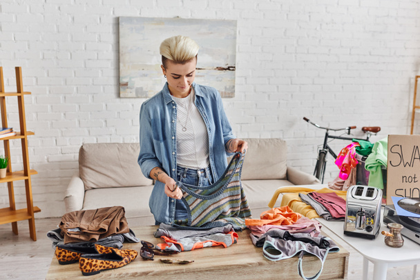 young and tattooed woman holding knitted tank top near wardrobe items, electric toaster and cezve while preparing pre-loved items for exchange, sustainable living and mindful consumerism concept - Photo, Image