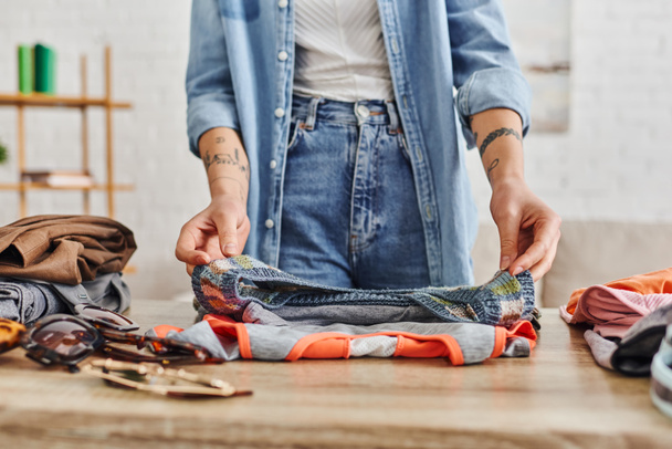 Teilbild einer jungen tätowierten Frau in Freizeitkleidung, die Kleiderschränke auf dem Wohnzimmertisch sortiert, Tauschbörse, umweltfreundliche Tauschgeschäfte, nachhaltiges Leben und bewusstes Konsumkonzept - Foto, Bild