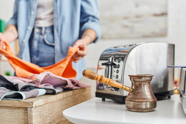 partial view of young woman sorting clothes near electric toaster and cezve, eco-friendly swaps, exchange market, blurred background, sustainable living and promoting circular economy concept - Photo, Image