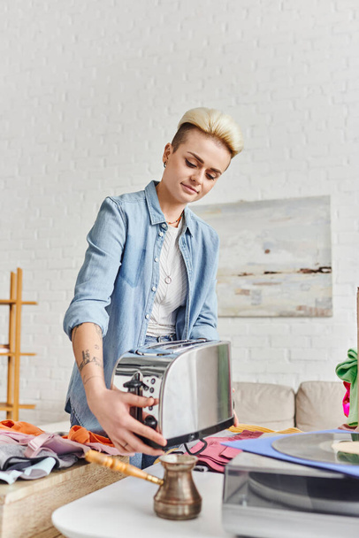 collaborative consumption, swap, smiling tattooed woman in casual clothes holding electric toaster near cezve and pre-loved clothes at home, sustainable living and promoting circular economy concept - Photo, Image