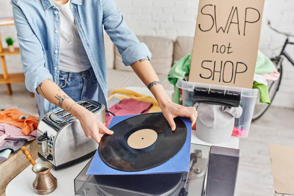 oříznutý pohled na tetovanou ženu držící vinylový disk poblíž gramofonu, elektrický toustovač, leštidlo, plastový obal s oblečením a výměnný lístek, koncept udržitelného bydlení a kruhového hospodářství - Fotografie, Obrázek
