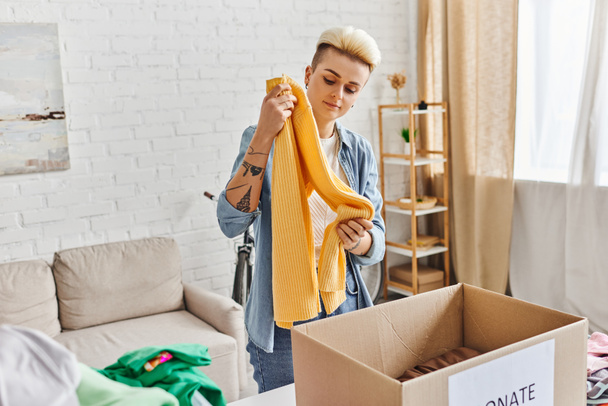 wolontariat i działalność charytatywna, modna i wytatuowana kobieta stojąca z żółtym swetrem w pobliżu pudełka tekturowego podczas sortowania odzieży do darowizn w domu, koncepcja zrównoważonego życia i odpowiedzialności społecznej - Zdjęcie, obraz