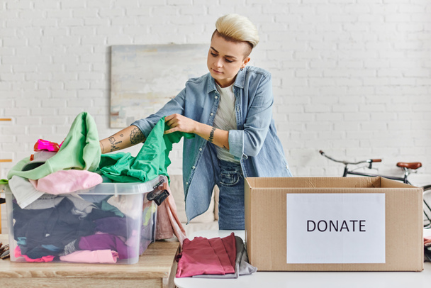 promoting social good, young tattooed woman sorting clothing in plastic container near carton box with donate lettering in modern living room, sustainable living and social responsibility concept - Photo, Image