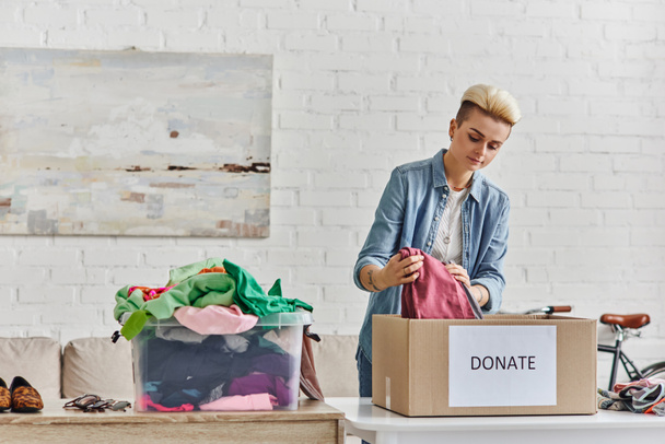 Bir amaç için bağış yapmak, şık dövmeli bir kadın plastik kutunun yanında giysilerini paketliyor oturma odasında ikinci el eşyalar, sürdürülebilir yaşam ve sosyal sorumluluk kavramı - Fotoğraf, Görsel