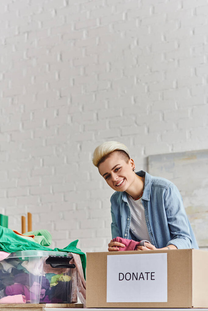 young and joyful woman with trendy hairstyle holding pre-loved clothes and looking at camera near plastic container and donation box at home, sustainable living and social responsibility concept - Photo, Image