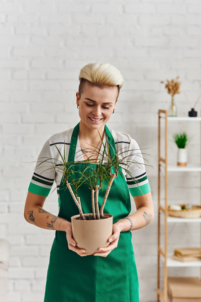overjoyed tattooed woman with trendy hairstyle holding potted exotic plant while standing in green apron in modern living room, plant therapy, sustainable home decor and green living concept - Photo, Image