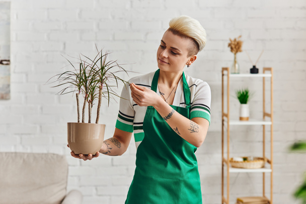 indoor gardening, young and positive tattooed woman in green apron touching leaves of tropical plant in modern apartment, plant care, sustainable home decor and green living concept - Photo, Image