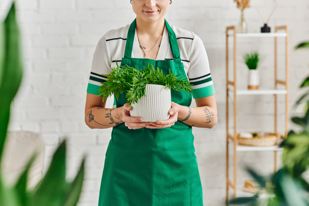 устойчивый дом, садоводство в помещении, частичный вид молодой татуированной женщины в фартуке держа цветочный горшок с зеленым растением и улыбаясь в современной квартире, зеленая концепция жизни - Фото, изображение
