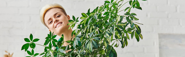 πορτρέτο της νεαρής γυναίκας με χαρούμενο χαμόγελο και μοντέρνο χτένισμα κοιτάζοντας κάμερα κοντά στο πράσινο houseplant στο σαλόνι, εσωτερική κηπουρική, βιώσιμη διακόσμηση σπιτιού και πράσινο έννοια διαβίωσης, πανό - Φωτογραφία, εικόνα