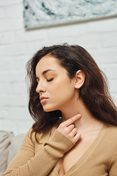 Young brunette woman in casual clothes closing eye while doing self-massage of thyroid gland on neck and promoting lymph flow at home, self-care ritual and holistic wellness practices concept - Photo, Image