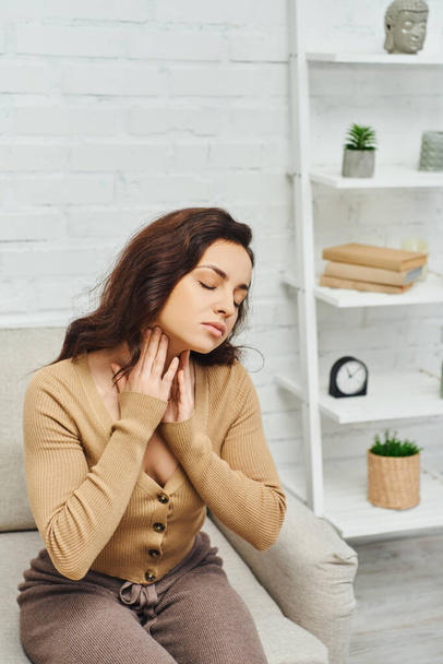 Brunette woman in casual clothes massaging lymphatic nodes on neck while doing therapeutic self-massage and sitting on couch at home, self-care ritual and holistic wellness practices concept - Photo, Image