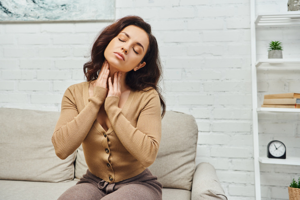 Young brunette woman in jumper closing eyes while massaging lymphatic nodes on neck during self-massage and sitting on couch at home, self-care ritual and holistic wellness practices concept - Photo, Image