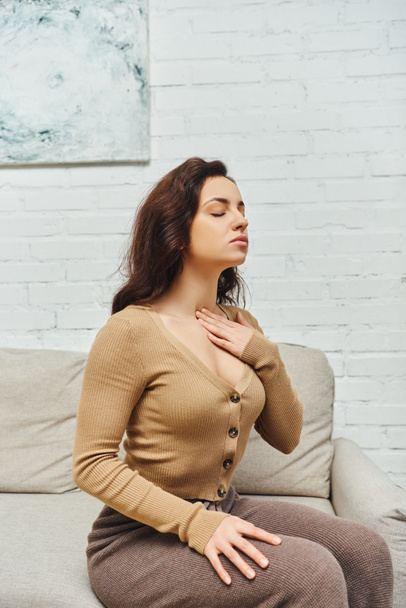 Young brunette woman in jumper touching neck while doing lymphatic nodes massage for drainage and support and sitting on couch at home, self-care ritual and holistic wellness practices concept - Photo, Image