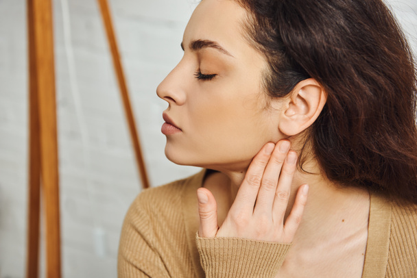 Side view of young brunette woman in brown jumper massaging face for lymphatic system support and drainage at home, self-care ritual and holistic healing concept, tension relief - Photo, Image