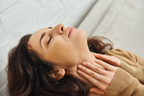 Close up view of relaxed brunette woman in brown jumper massaging neck during lymphatic drainage support and sitting on couch at home, self-care ritual and holistic healing concept, tension relief - Photo, Image