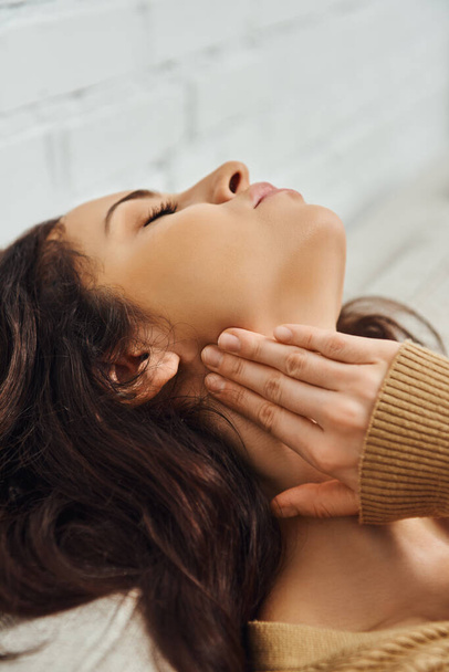 Jeune femme brune en pull brun touchant le nœud lymphatique sur le cou et se relaxant pendant l'auto-massage sur le canapé à la maison, rituel d'auto-soin et concept de guérison holistique, soulagement de la tension - Photo, image