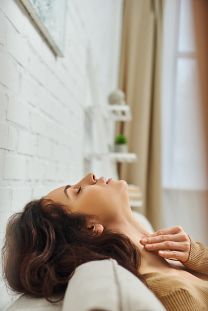 Side view of young brunette woman in brown jumper massaging thyroid gland and neck while relaxing and sitting on couch at home, self-care ritual and holistic healing concept, tension relief - Photo, Image