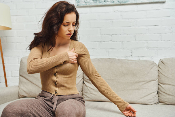 Young brunette woman in brown jumper suffering from pain while massaging lymphatic nodes on armpit and sitting on couch at home, self-care ritual and holistic wellness practices concept - Photo, Image