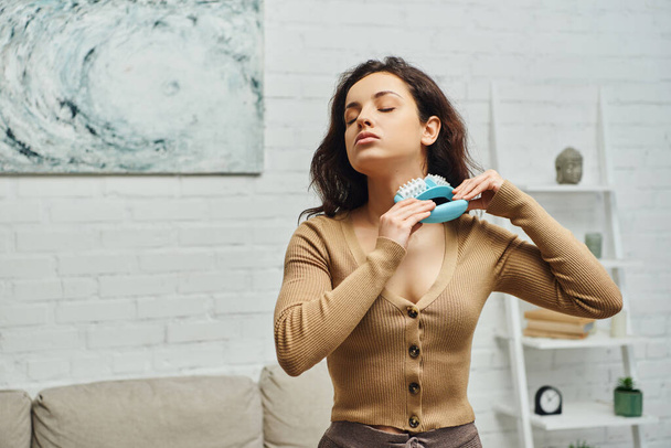 Junge brünette Frau im braunen Pullover massiert Lymphsystem am Nacken mit Massagegerät im Stehen im modernen Wohnzimmer zu Hause, Selbstversorgungsritual und ganzheitliches Wellness-Praxiskonzept - Foto, Bild