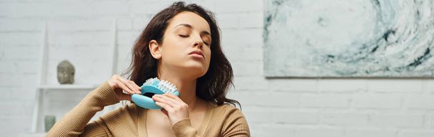 Relaxed young brunette woman in casual jumper massaging neck with massager during self-massage of lymphatic system in living room, self-care ritual and holistic wellness and health concept, banner - Photo, Image
