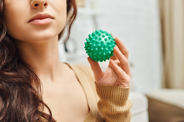 Cropped view of young brunette woman in jumper holding manual massage ball while standing in blurred living room at home, lymphatic system support and home-based massage, tension relief - Photo, Image