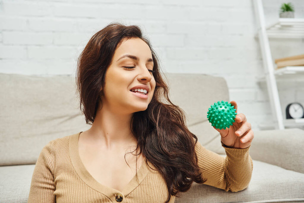 Cheerful young brunette woman in casual jumper looking at manual massage ball while sitting near couch at home, home-based massage and holistic wellness practices concept, balancing energy - Photo, Image